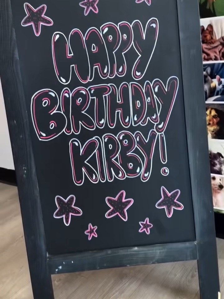 A chalkboard easel reading Happy Birthday Kirby in pink bubble letters with stars, in front of a wall of dog photographs.